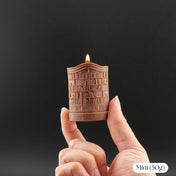 A hand holds a miniature brown wax candle shaped like a bookshelf, with a small flame burning from the top, against a dark gray background. A white label in the bottom right corner reads 'Mini (50g)'.