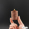 A hand holds a miniature brown wax candle shaped like a bookshelf, with a small flame burning from the top, against a dark gray background. A white label in the bottom right corner reads 'Mini (50g)'.