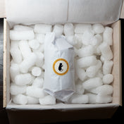 A white paper-wrapped package with a circular black cat logo and gold text sits centered inside an open cardboard box filled with white packing foam.