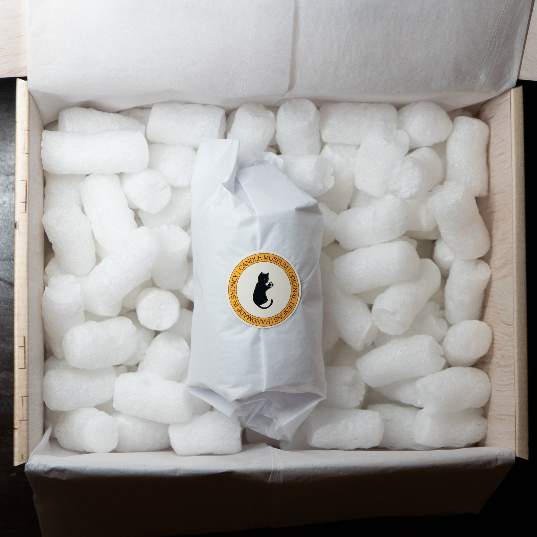 A white paper-wrapped package with a circular black cat logo and gold text sits centered inside an open cardboard box filled with white packing foam.
